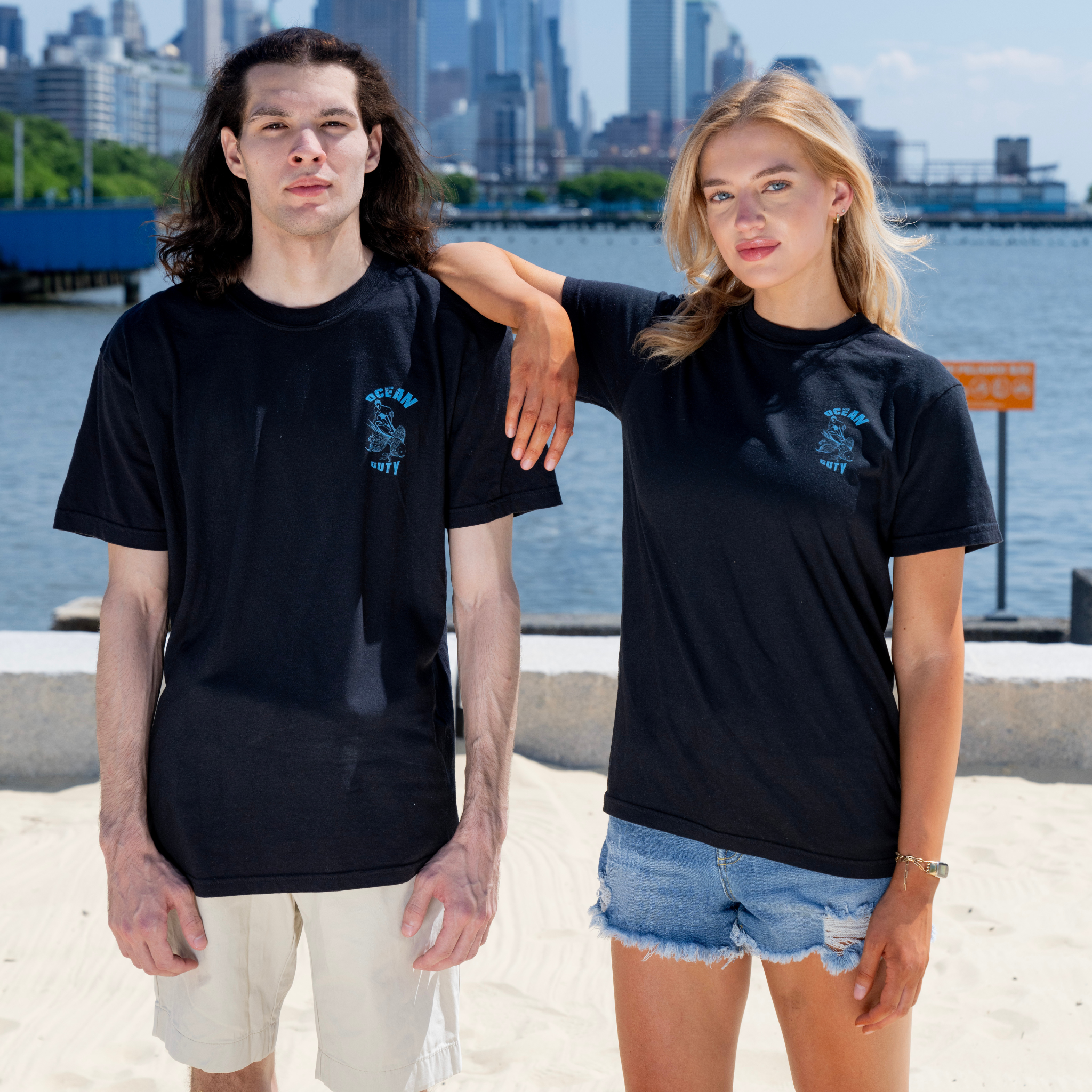 Two people wearing blue t-shirts at the beach with ocean themed graphic print on the upper left side chest.