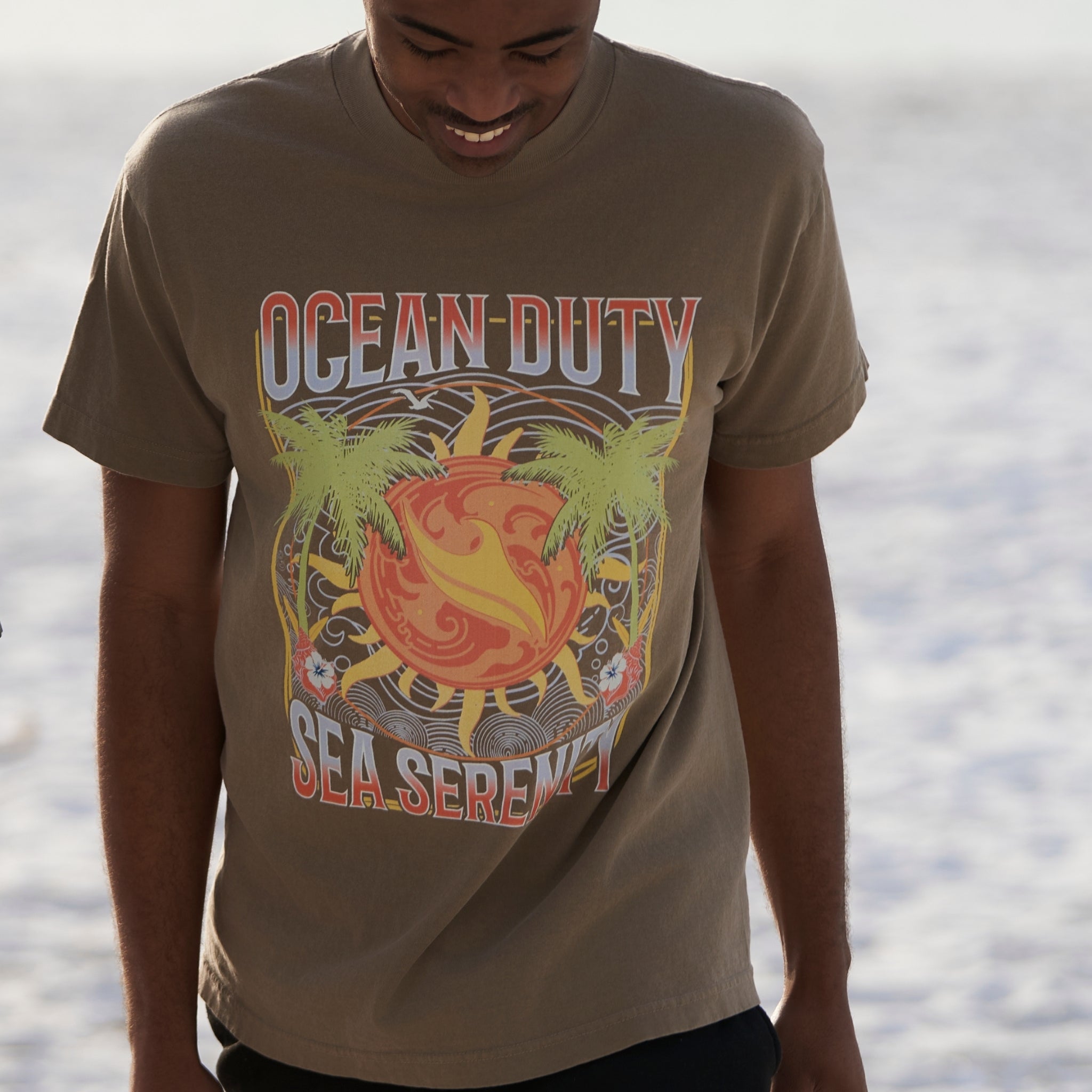 A male wearing a faded brown t-shirt with tropical designs walking on the beach.
