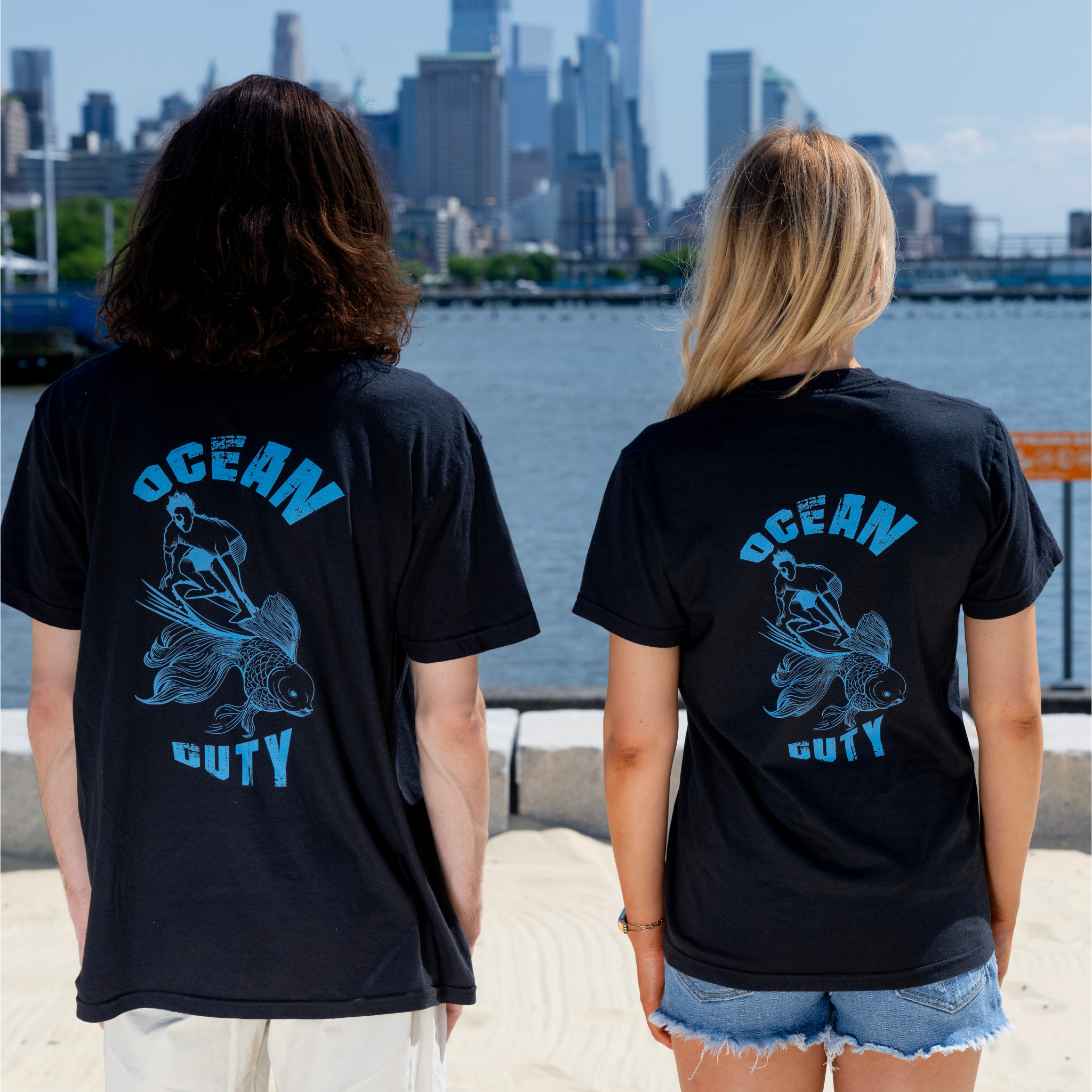 Two people wearing black t-shirts with a man surfing on a goldfish and ocean duty printed on it.