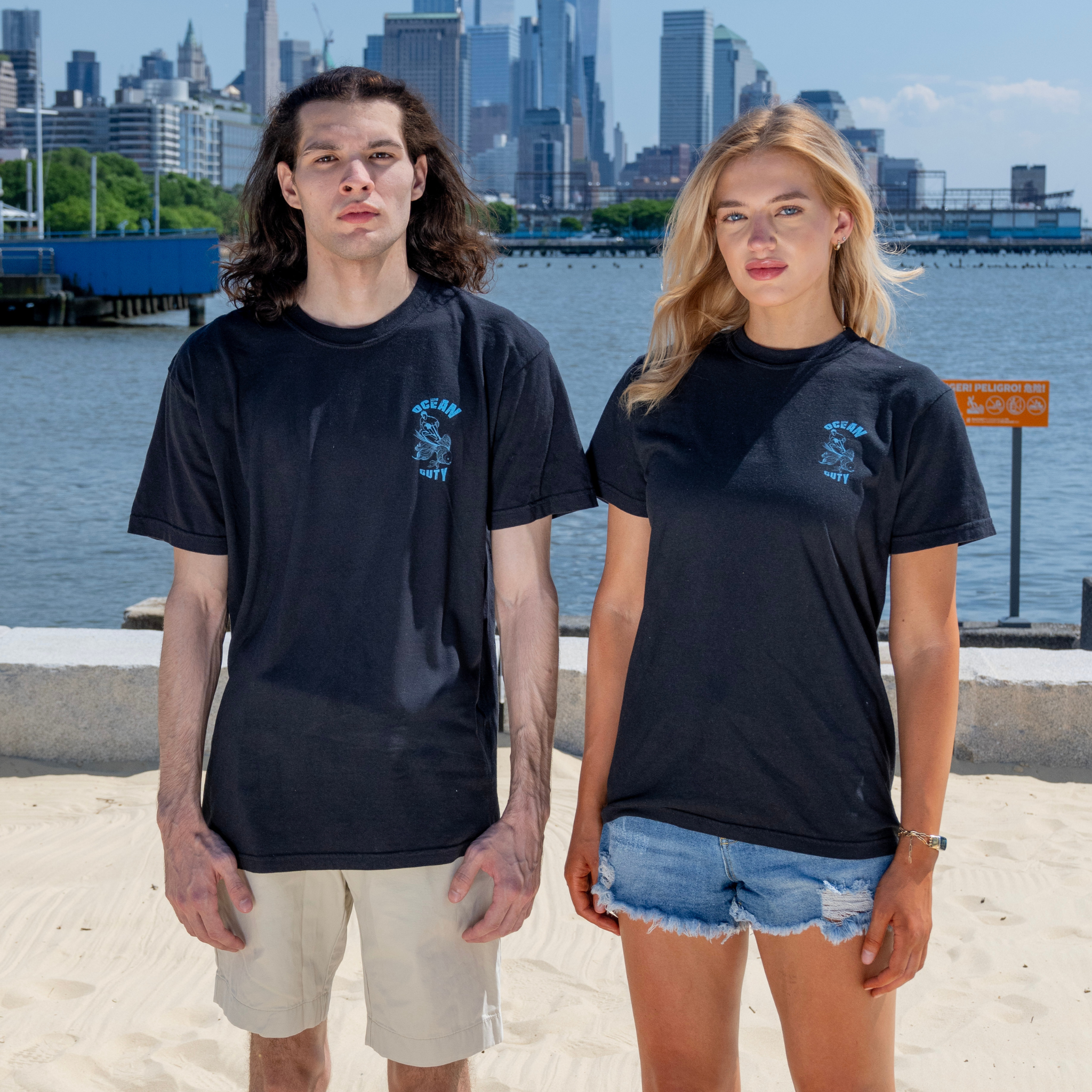 Two people wearing black t-shirts with a man surfing on a goldfish and ocean duty printed on it.