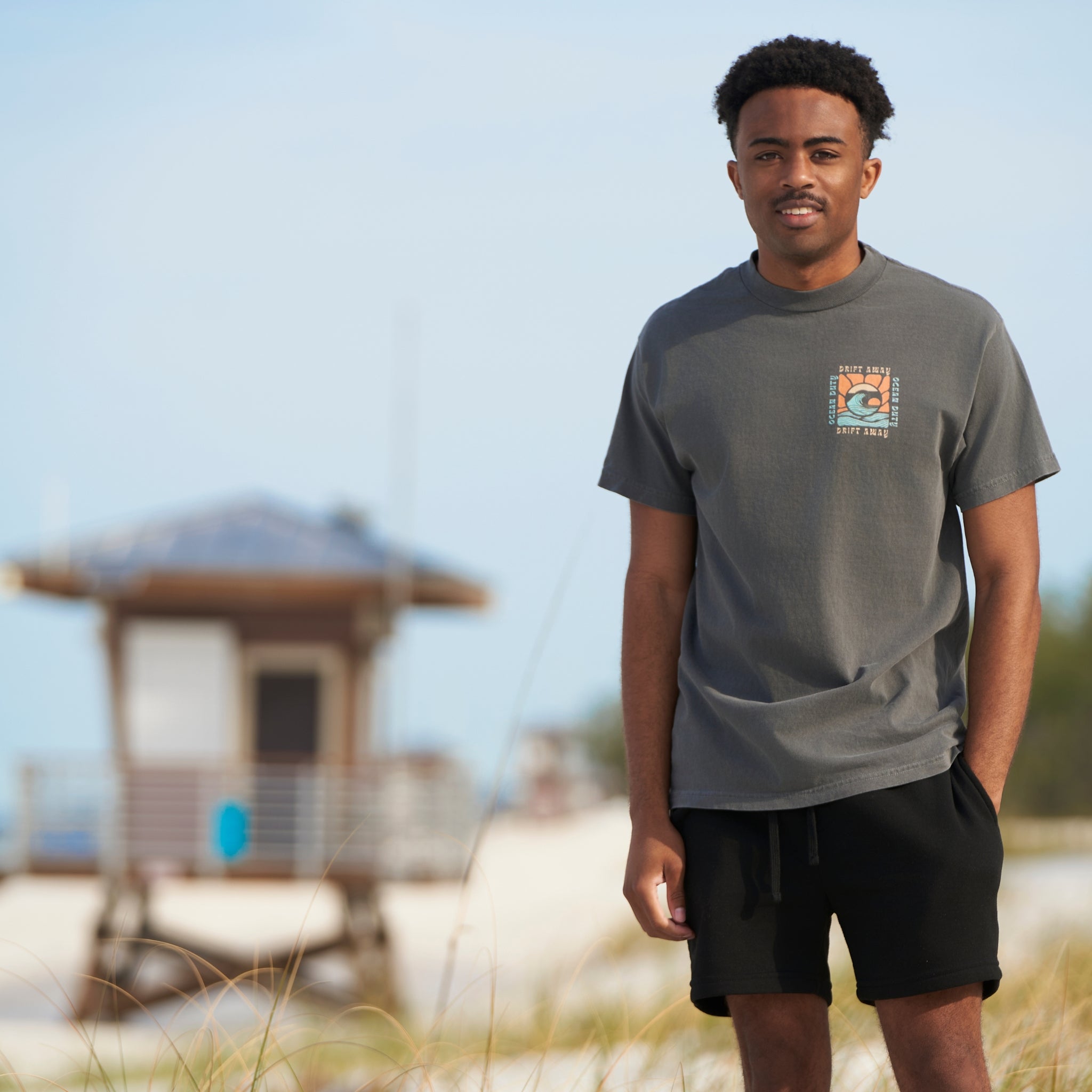 Man on the beach wearing a faded black t-shirt with graphic print of a ocean wave and the words 'drift away.'