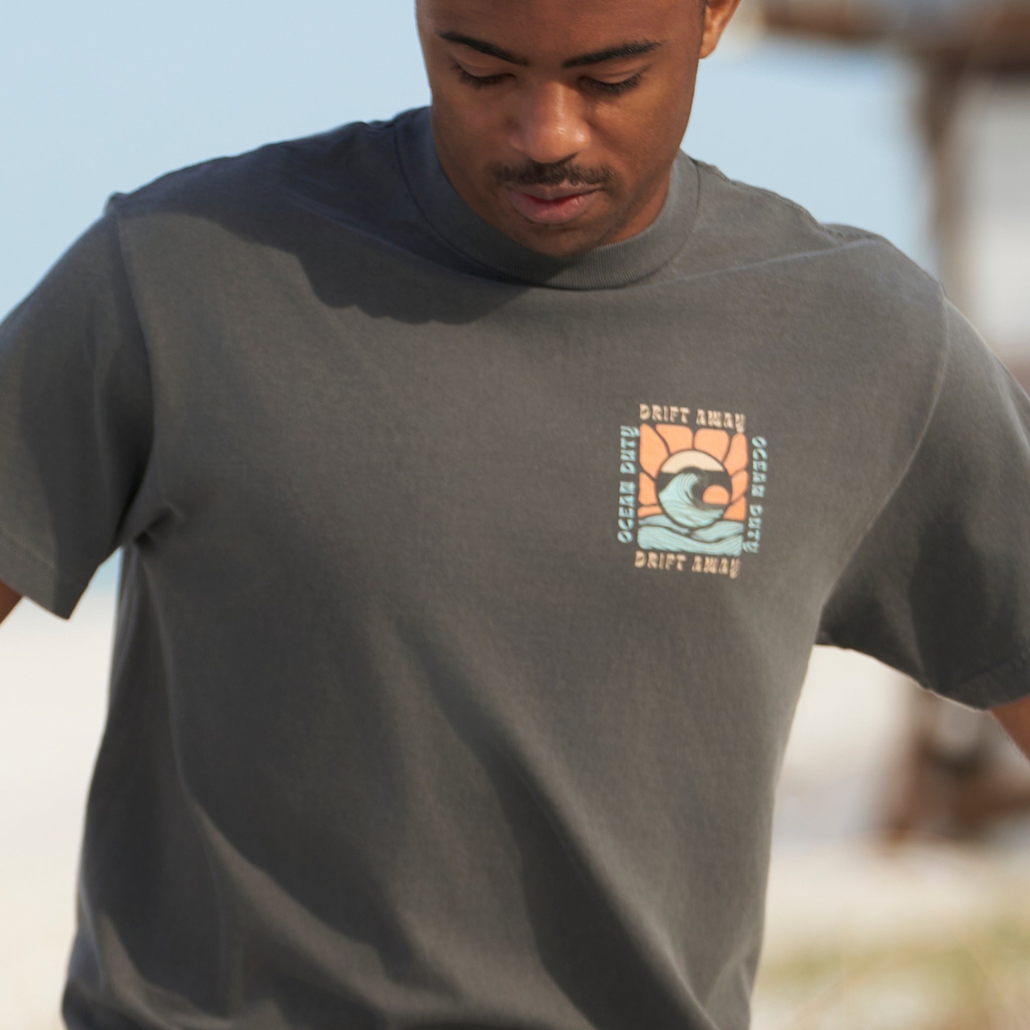 A man on the beach wearing a faded black t-shirt with graphic print of a ocean wave and the words 'drift away.'