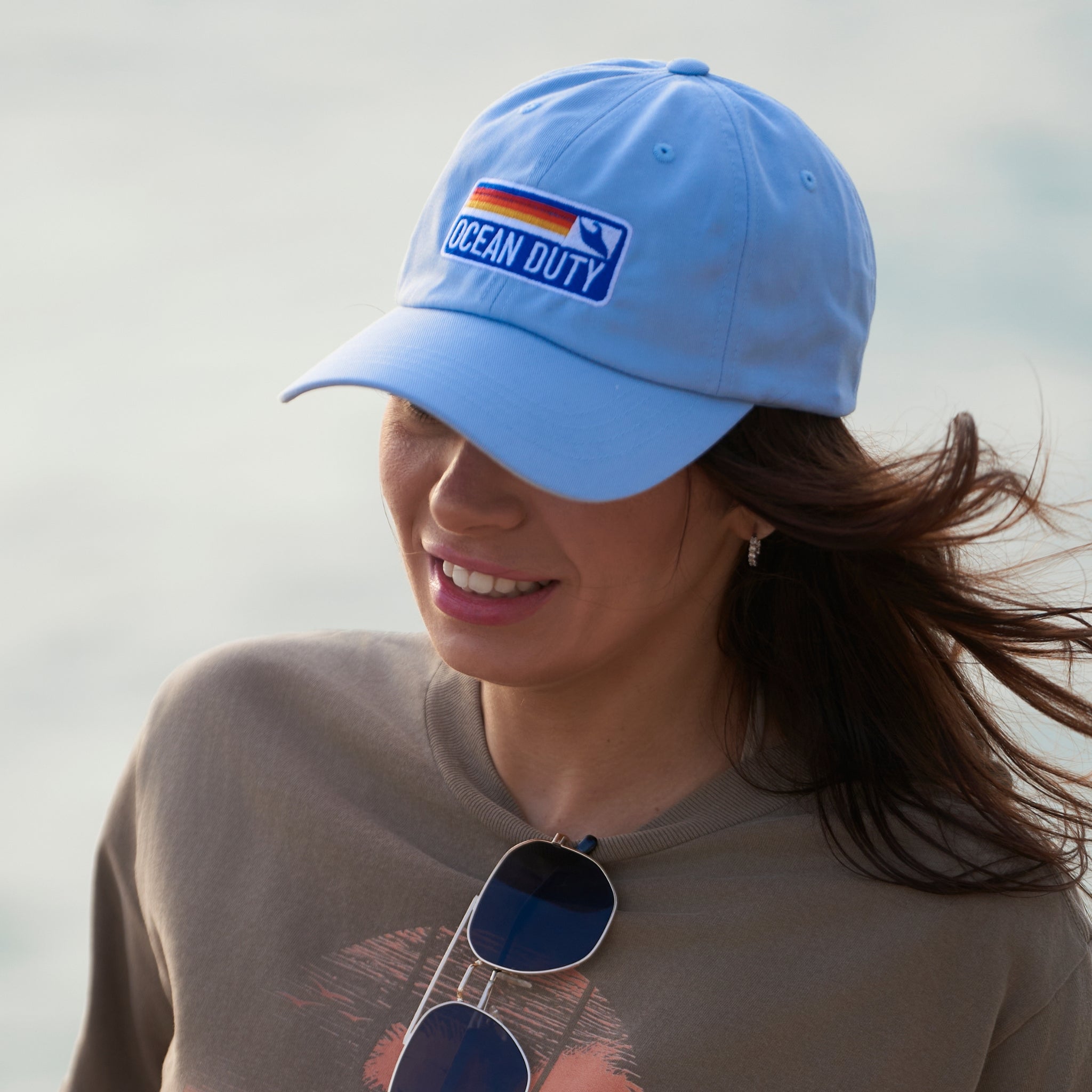 Woman wearing a a light blue beach style embroidered cotton hat with a adjustable strap. The embroidery has bright colored stripes and says 'Ocean Duty'.