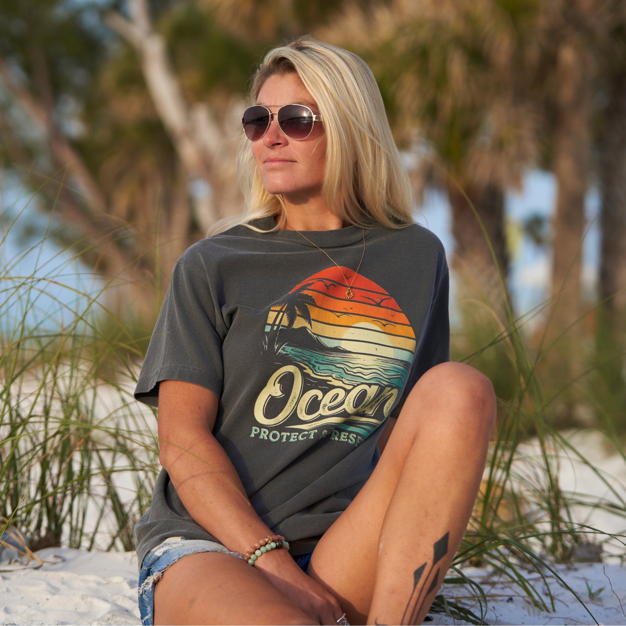 A woman sitting on the beach wearing a faded black t-shirt with an ocean sunset over a beach with the words 'protect & preserve' printed below.'