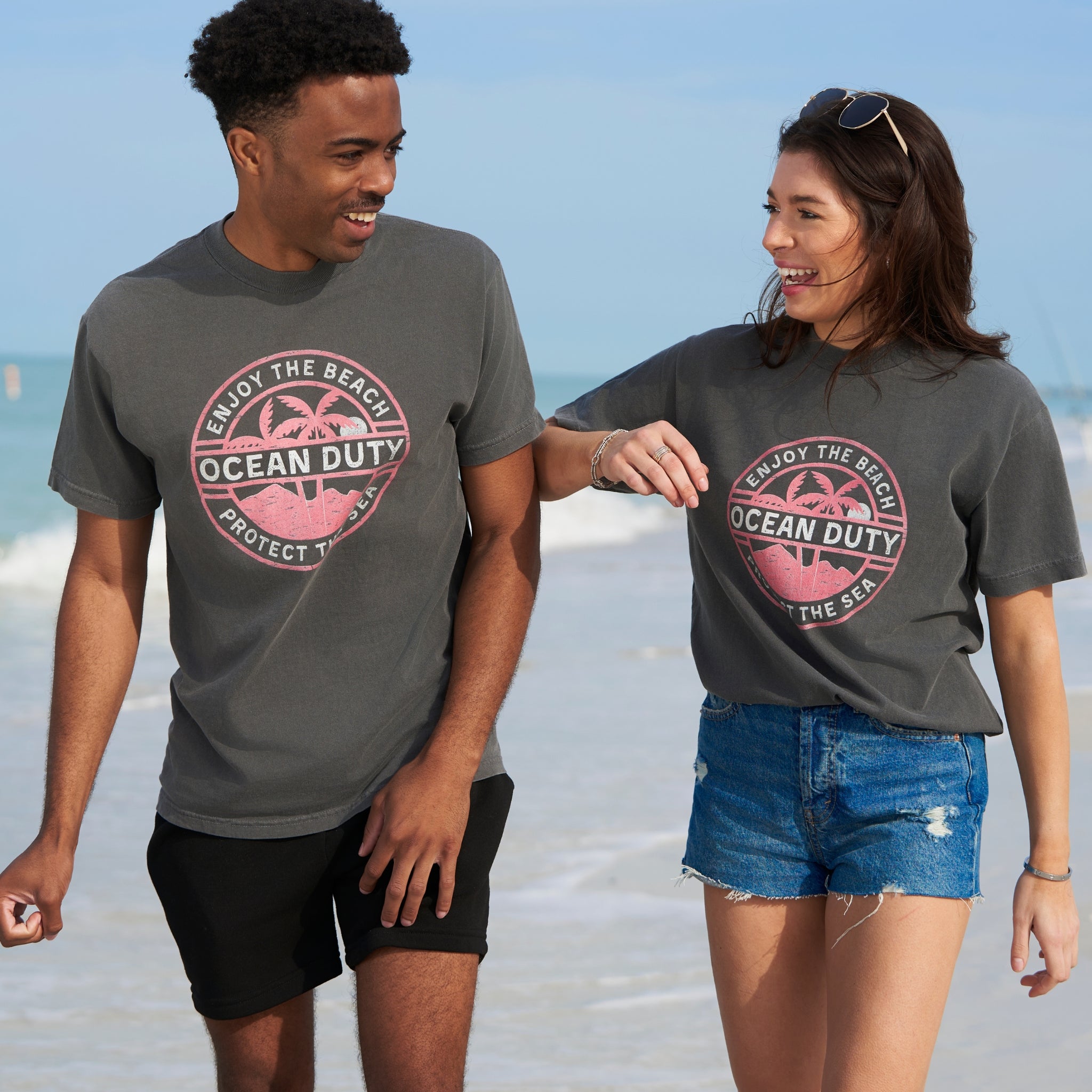 A male and female on the beach wearing faded black T-shirts with a red and white tropical beach graphic design printed on the front about ocean conservation.