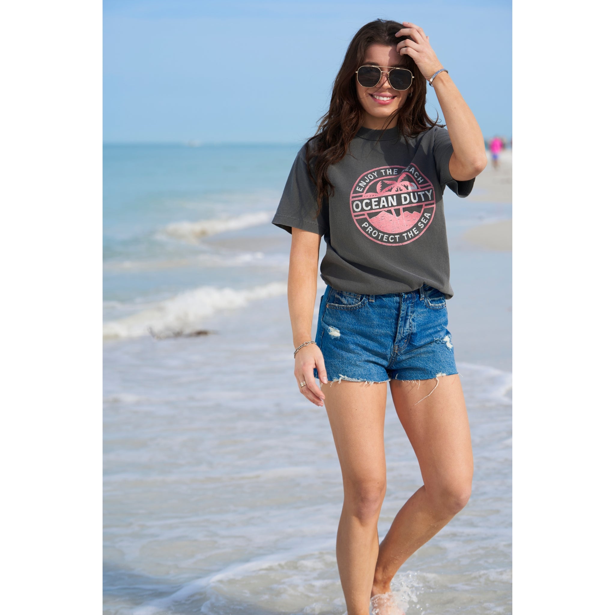 A female on the beach wearing a faded black T-shirt with a red and white tropical beach graphic design printed on the front about ocean conservation.