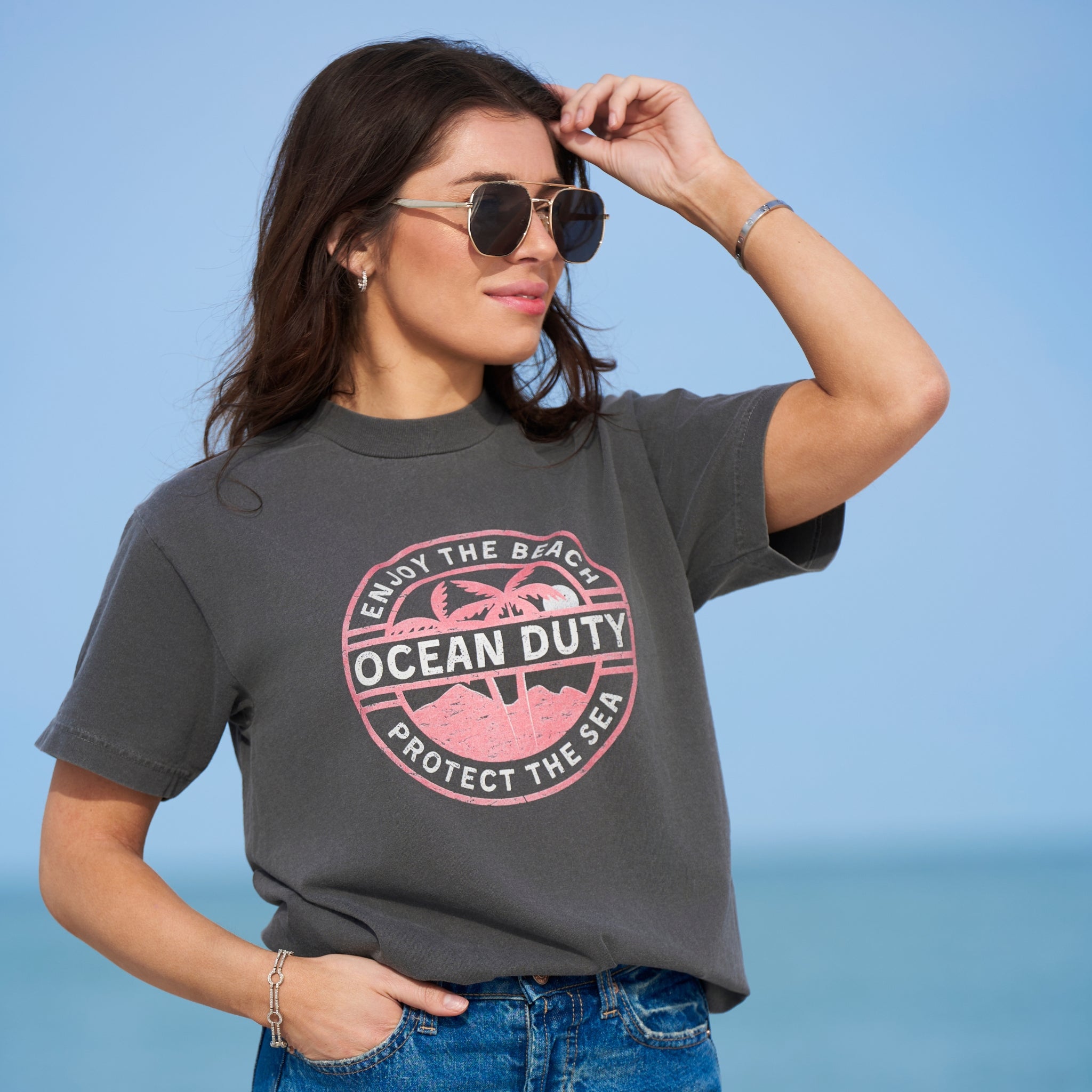 A female standing on the beach wearing  a faded black T-shirt with a red and white tropical beach graphic design printed on the front about ocean conservation.