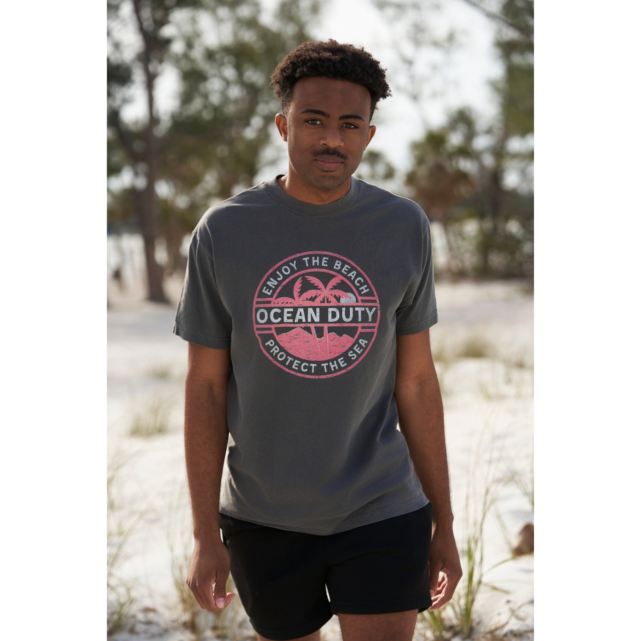 A male on the beach wearing a faded black T-shirt with a red and white tropical beach graphic design printed on the front about ocean conservation.