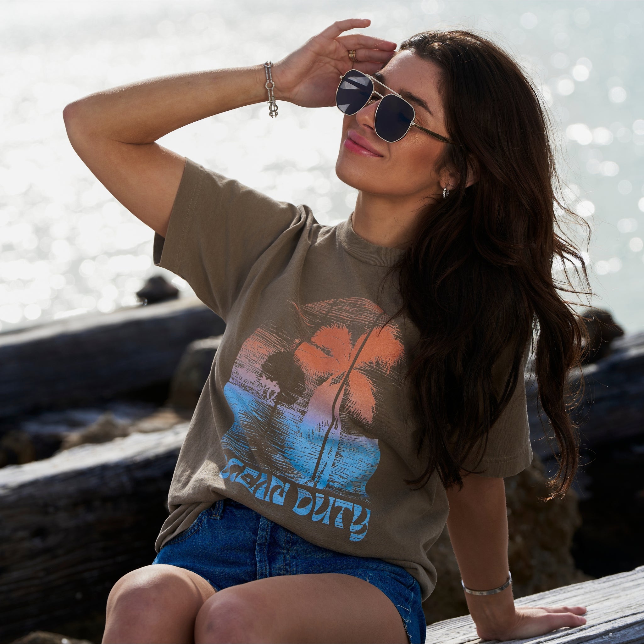 A woman sitting down wearing a faded brown T-shirt with distressed colorful beach print on the the front.