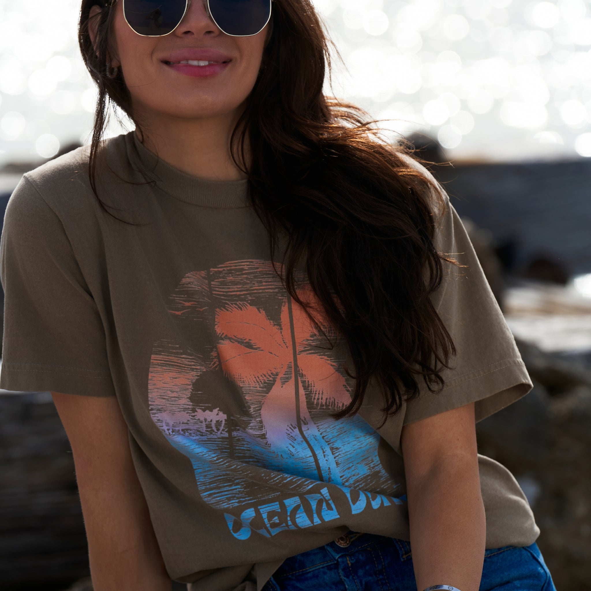 A woman on the the beach wearing a faded brown T-shirt with distressed colorful tropical print on the the front.