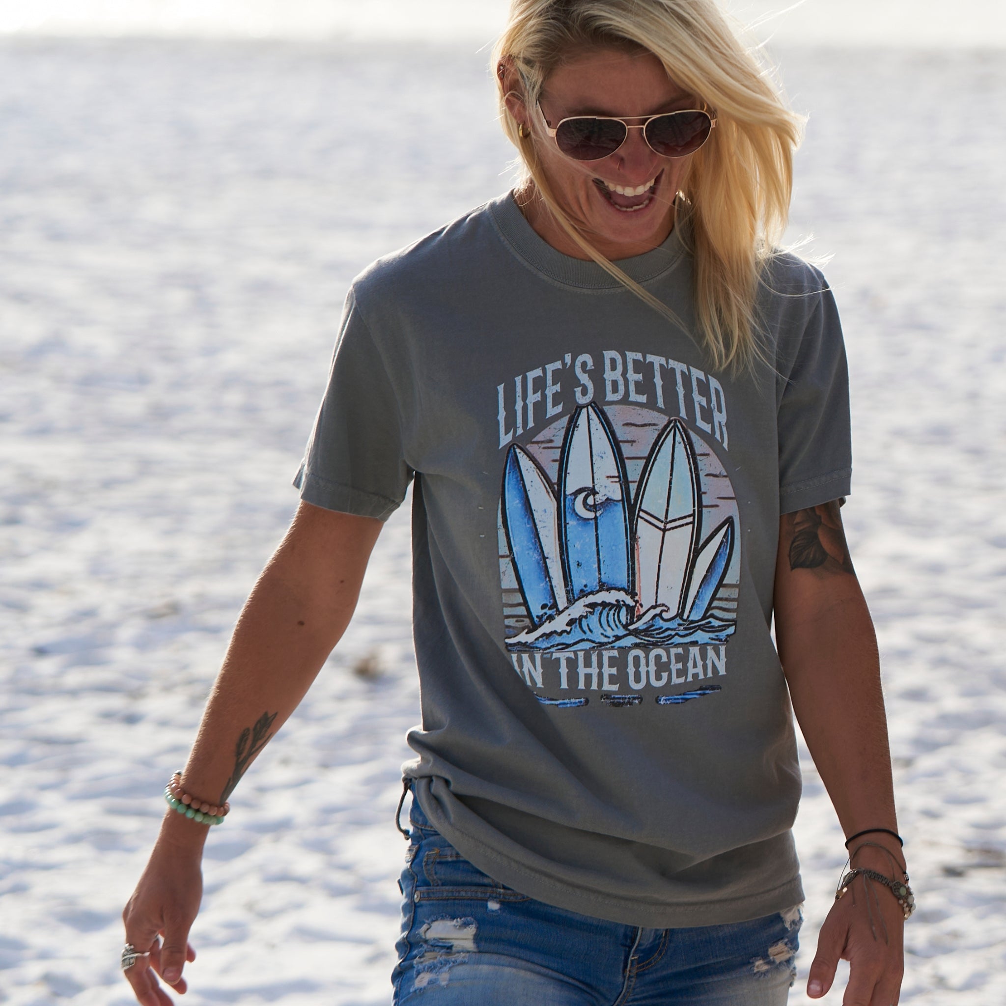 Woman at the beach wearing a grey T-shirt that has a graphical picture of surfboards on the ocean.