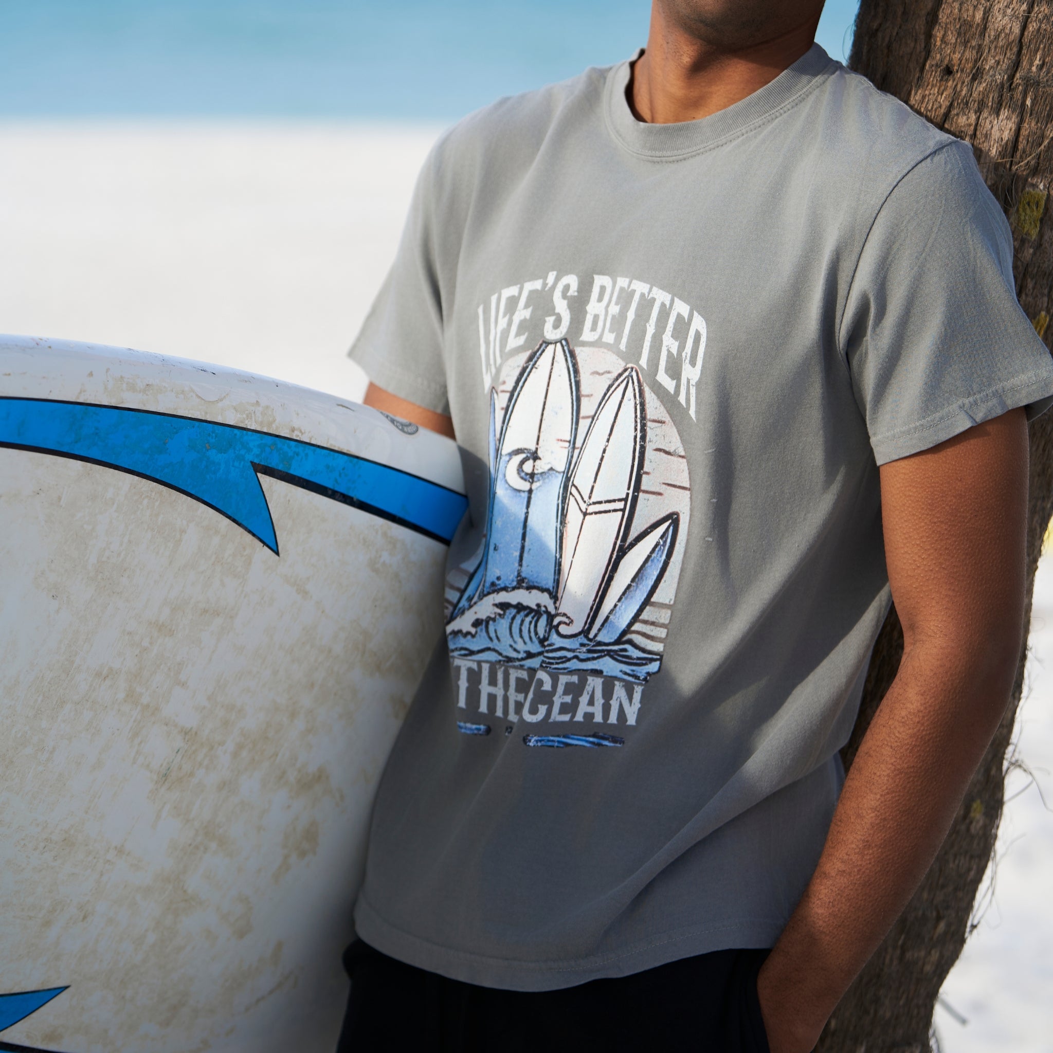 Man holding a surf board with a Grey T-shirt has a colorful graphic picture of surf boards at the ocean.