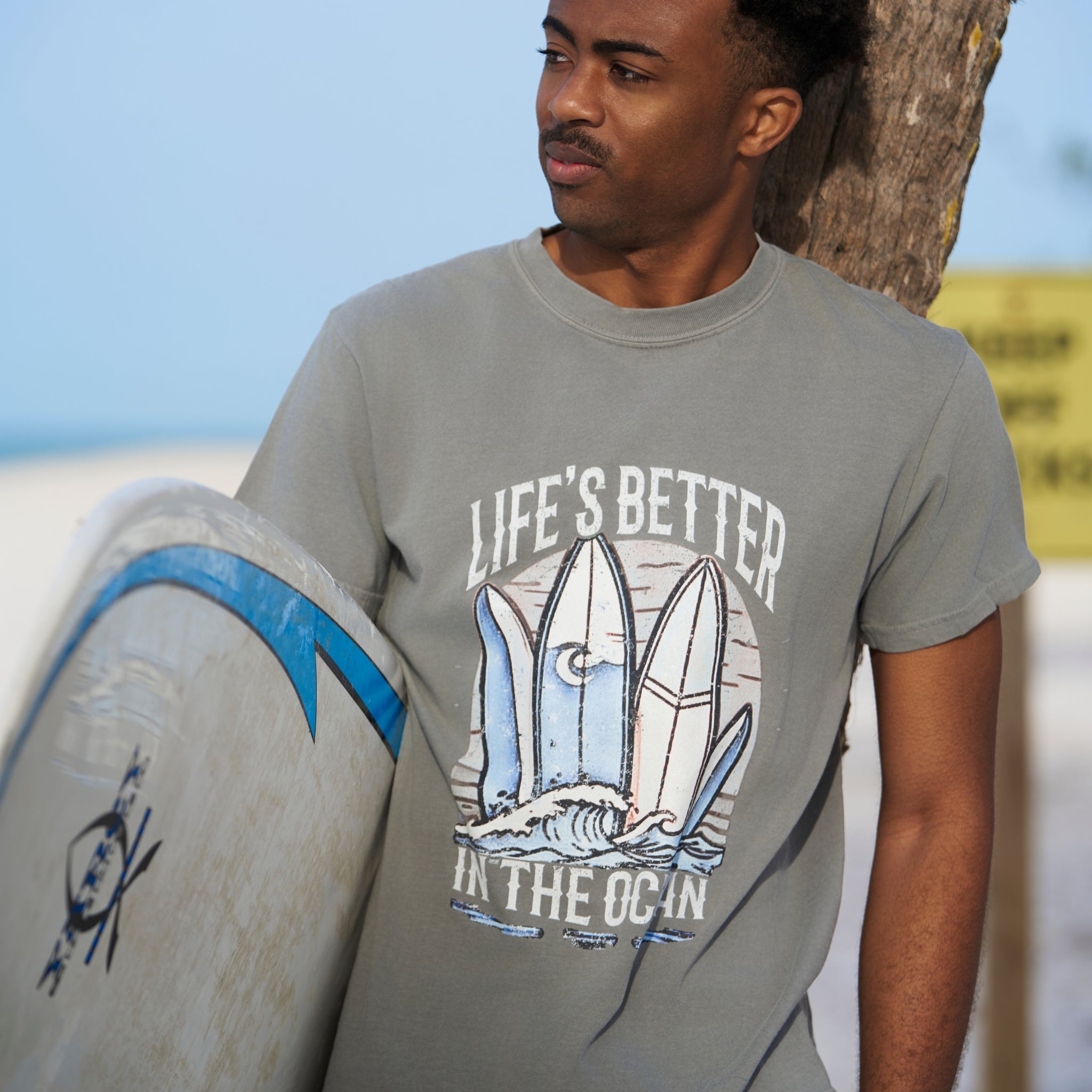 Man at a beach in wearing a Grey T-shirt that has a graphical picture of surfboards at the ocean.