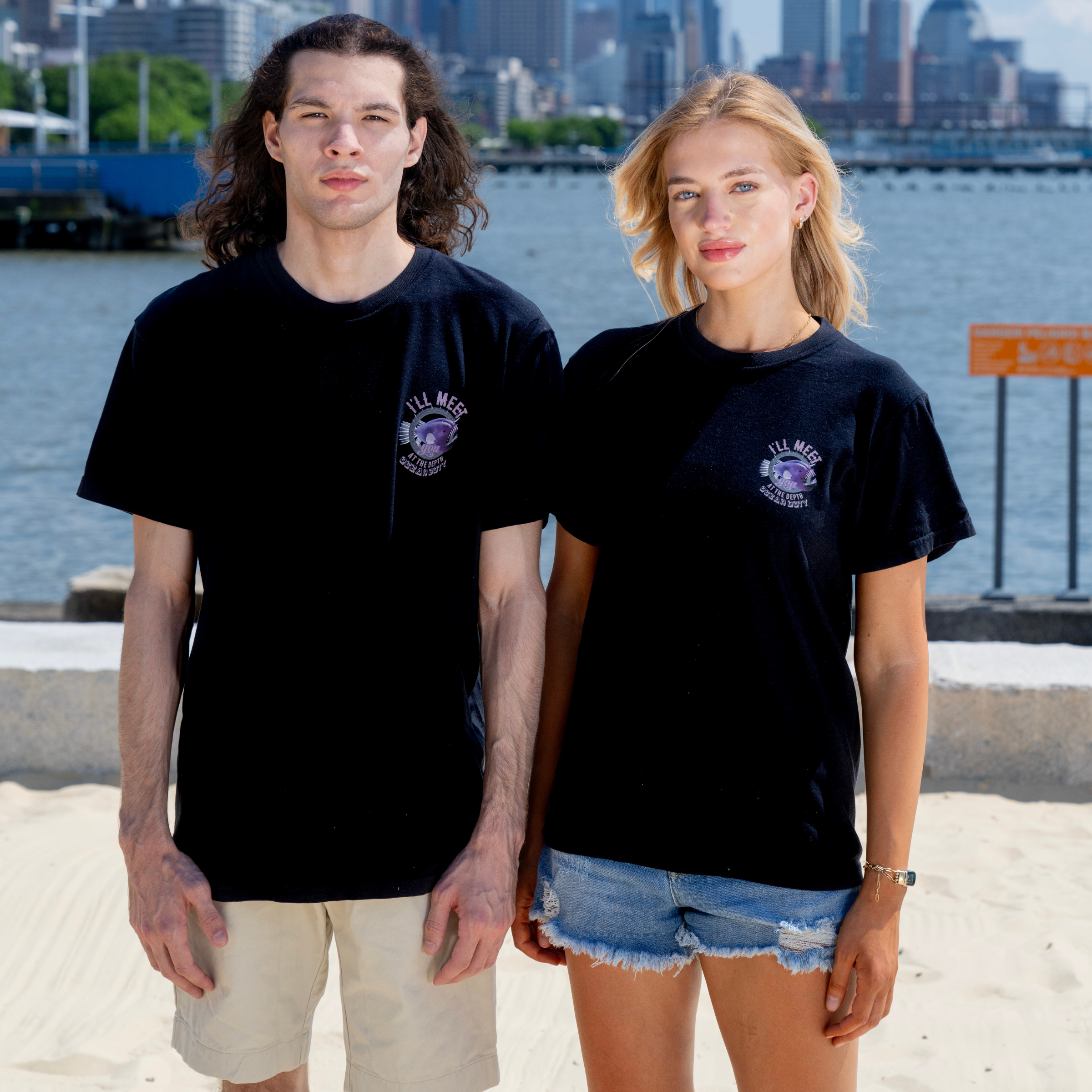A man and woman wearing a black T-shirt that represents ocean conservation, standing on the beach in front of the ocean. 