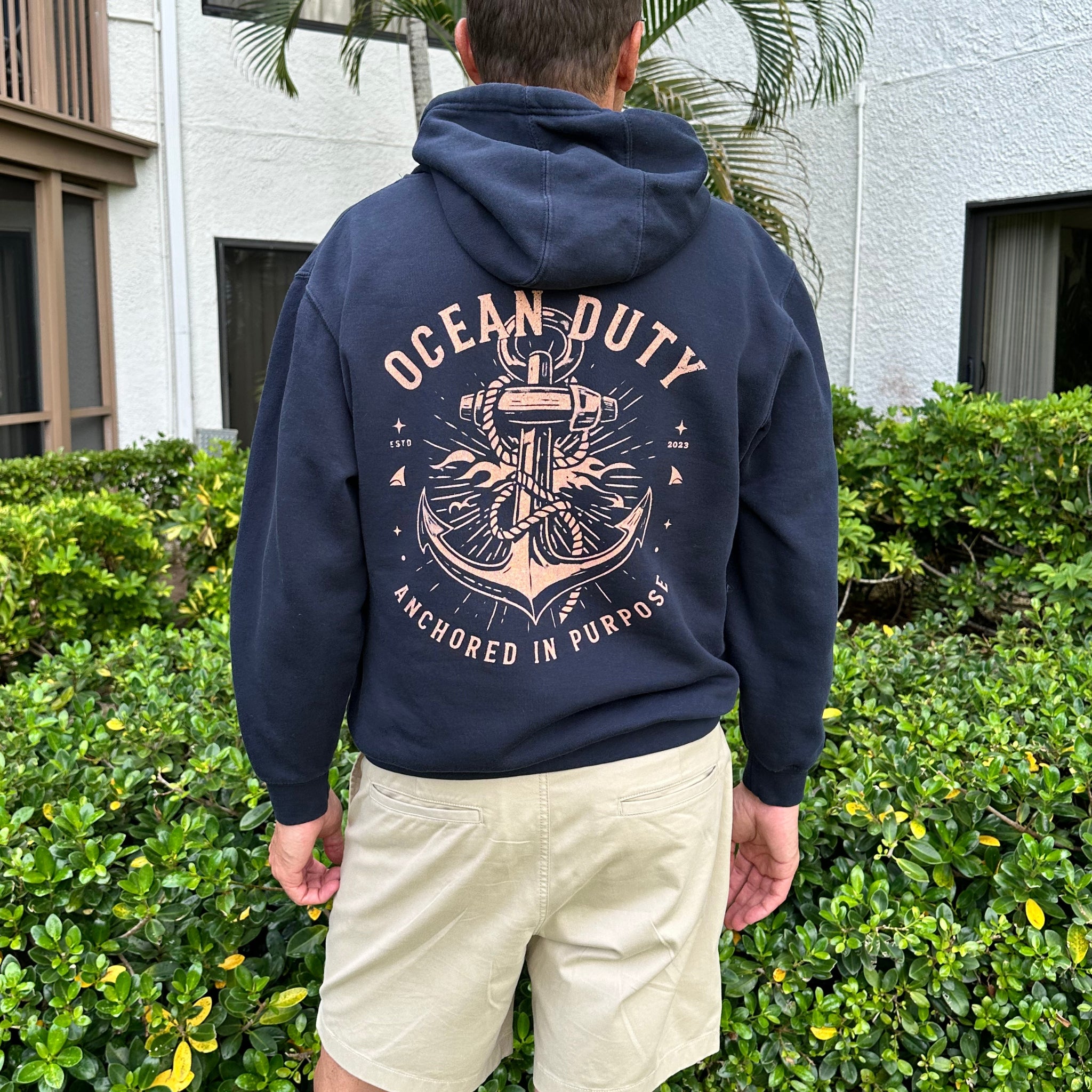 A man viewed from behind wearing a navy blue hoodie with a large anchor graphic and the words 'OCEAN DUTY' and 'ANCHORED IN PURPOSE' on the back, paired with light khaki shorts in an outdoor setting with green bushes and a building in the background.