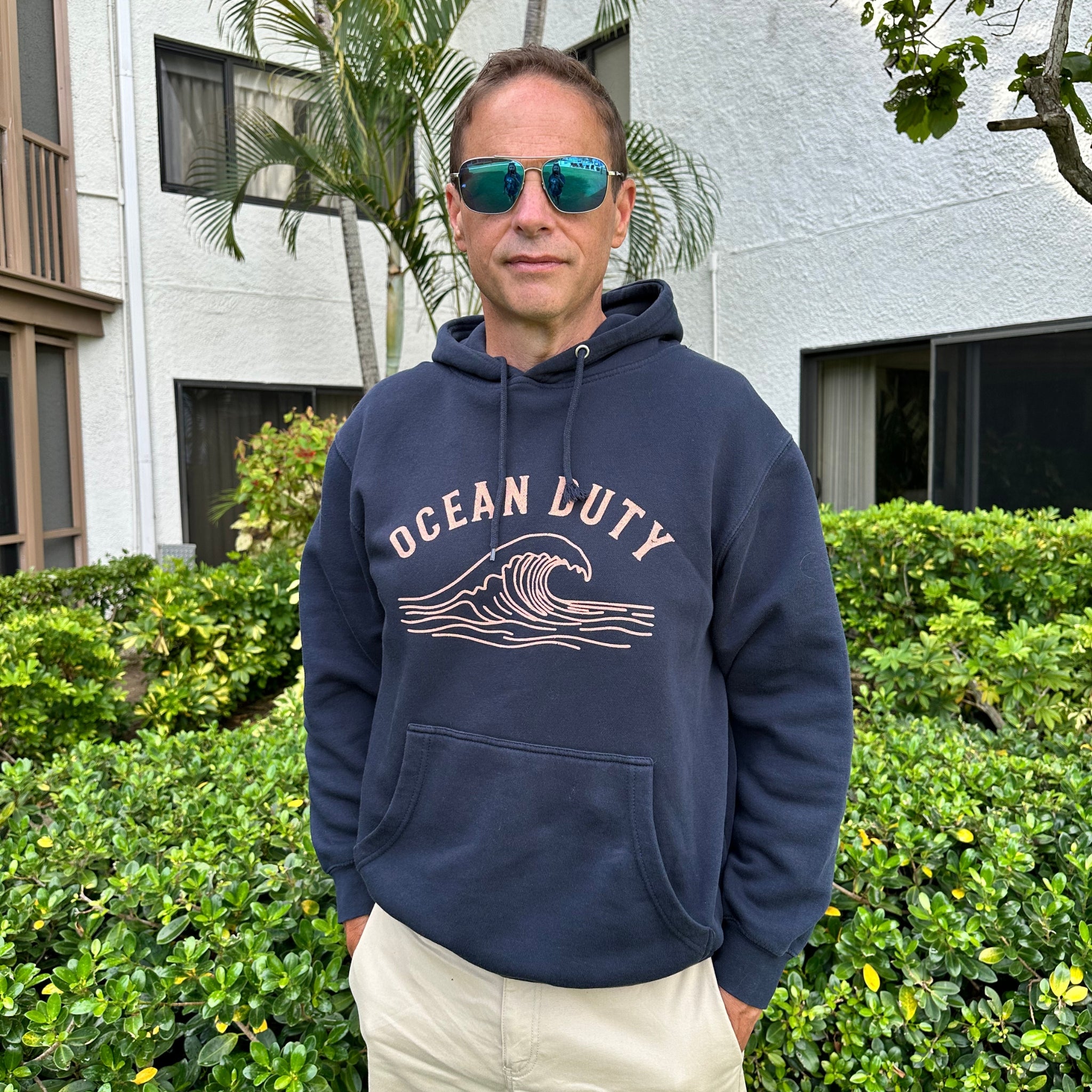 A man wearing a navy blue hoodie with a beige 'OCEAN LUTY' wave graphic on the front, standing outdoors in front of green hedges and a building.