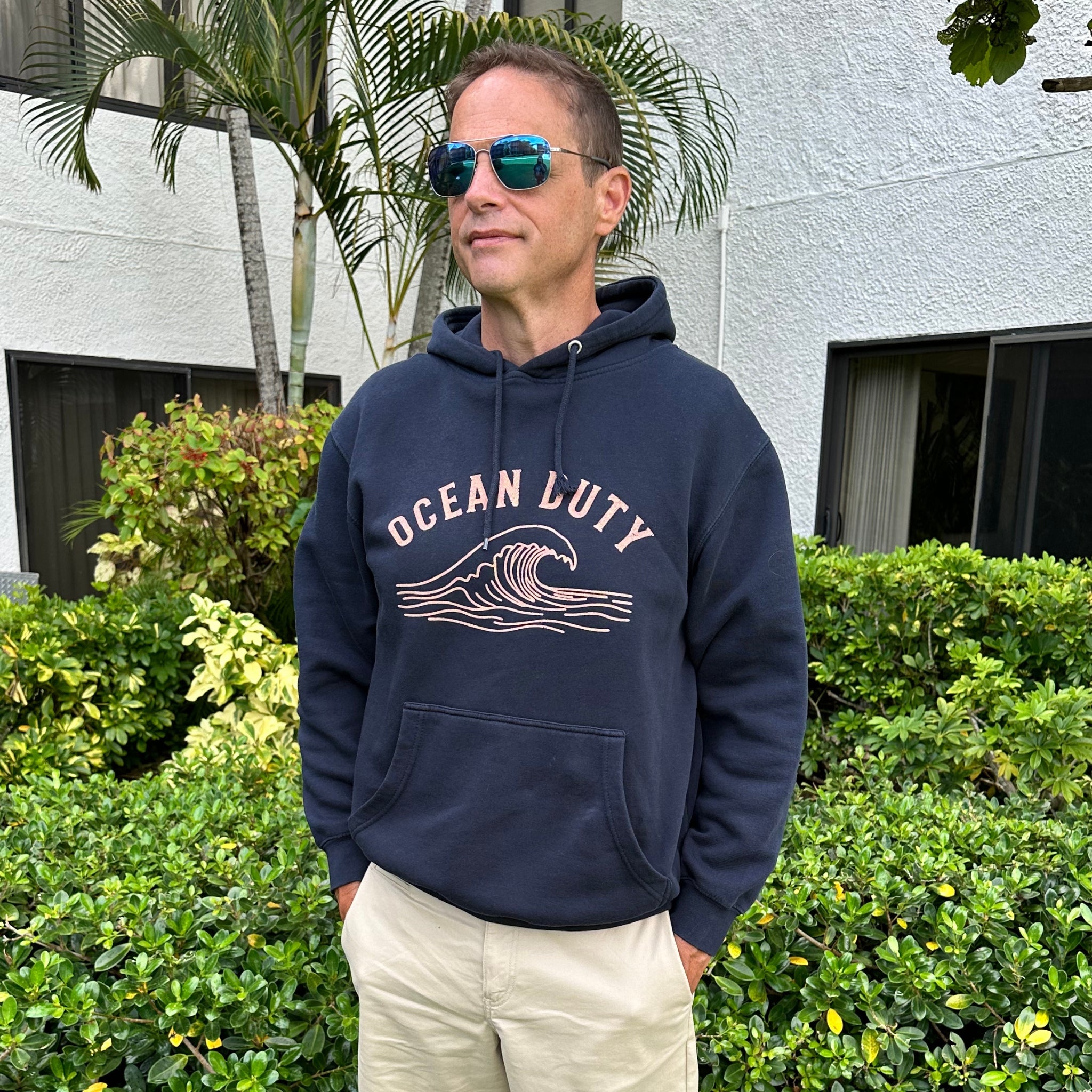 A man wearing a navy blue hoodie with 'OCEAN LUTY' and a wave graphic printed in beige on the front, paired with khaki pants and reflective sunglasses, standing outdoors in front of a white building and green bushes.