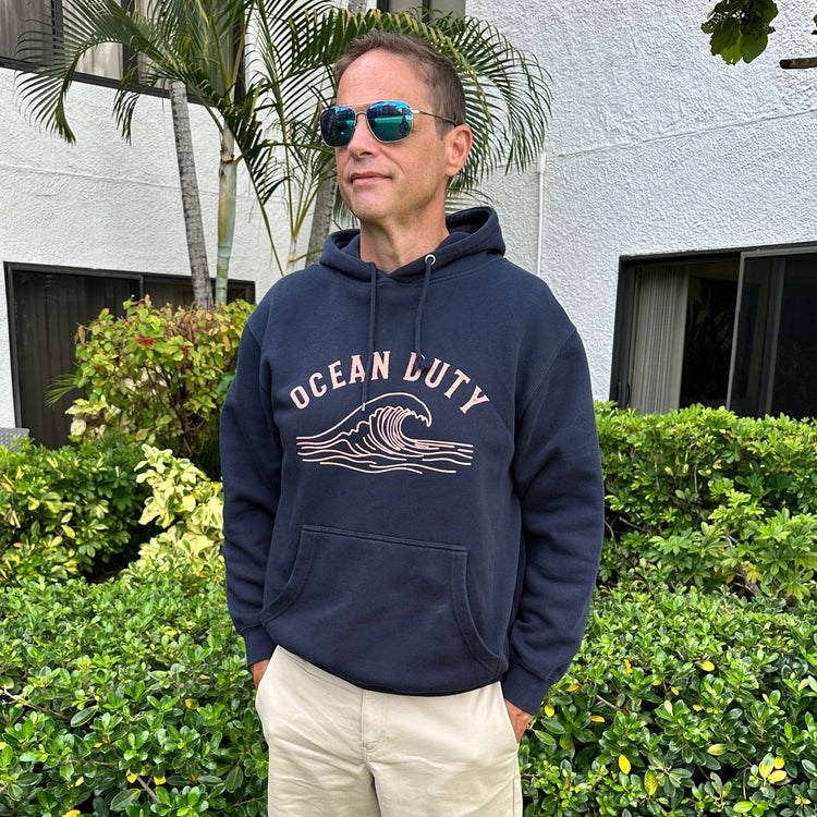 A man wearing a navy blue hoodie with 'OCEAN LUTY' and a wave graphic printed in beige on the front, paired with khaki pants and reflective sunglasses, standing outdoors in front of a white building and green bushes.