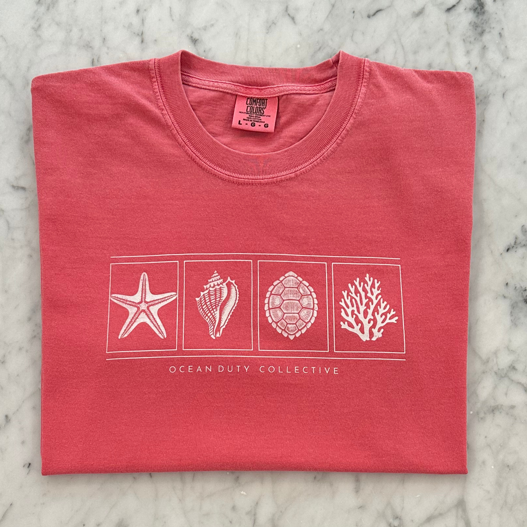 A folded coral pink t-shirt with a white graphic print of seashells and marine life on a marble background. The brand 'Ocean Duty Collective' is printed below the graphic.
