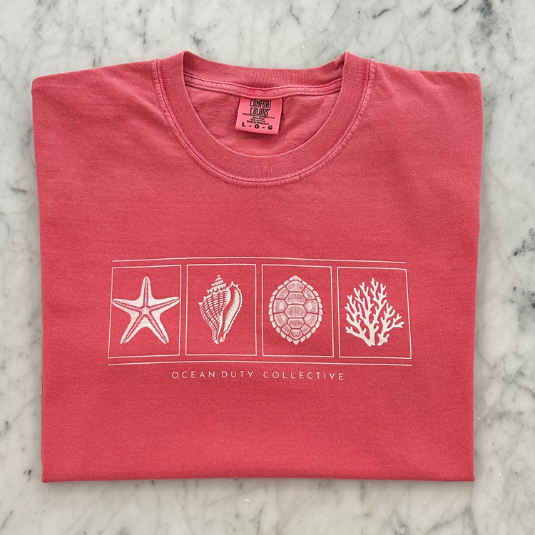 A folded coral pink t-shirt with a white graphic print of seashells and marine life on a marble background. The brand 'Ocean Duty Collective' is printed below the graphic.
