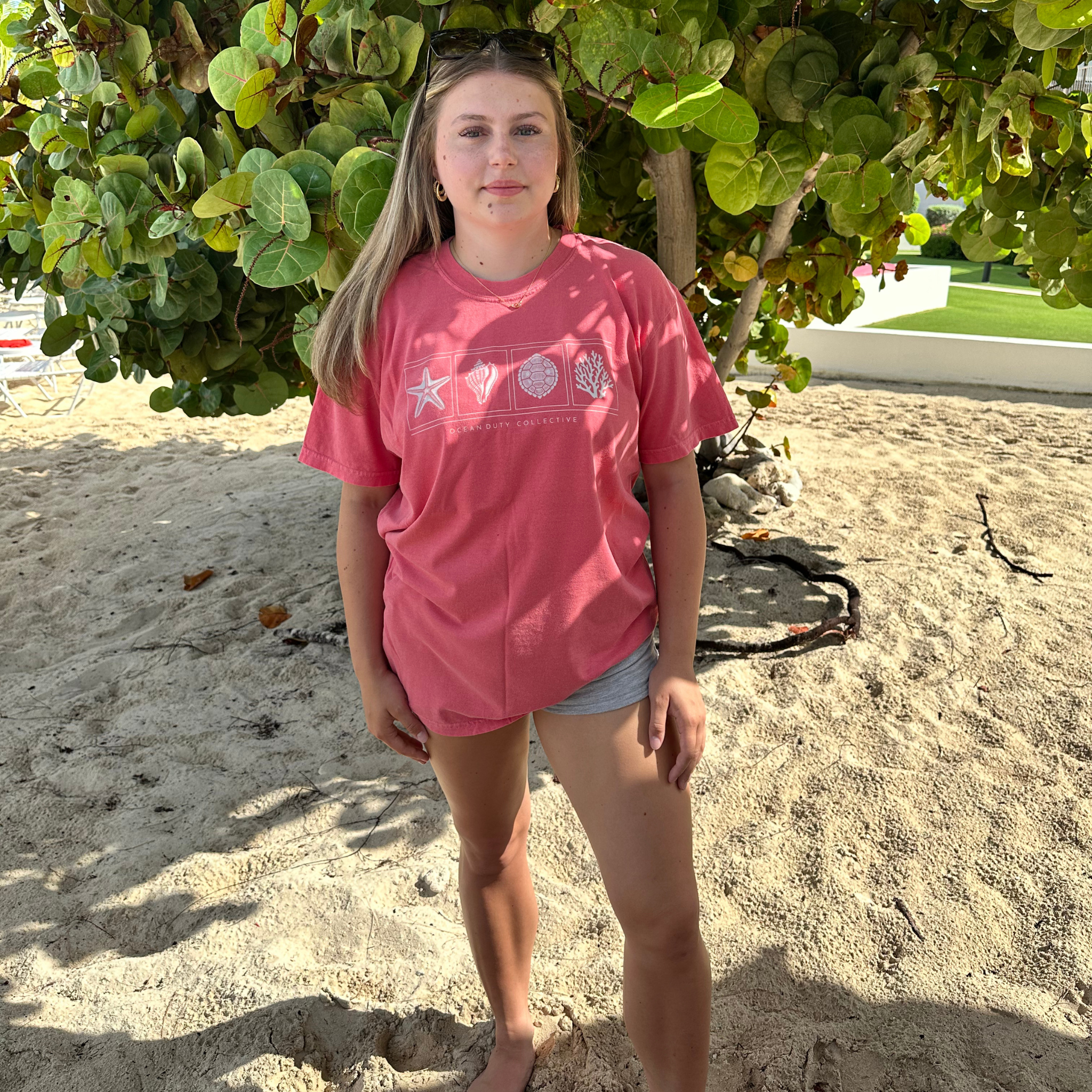 A woman standing on a sandy beach under a leafy tree, wearing a pink graphic t-shirt with white seashell and starfish designs, and light gray denim shorts.