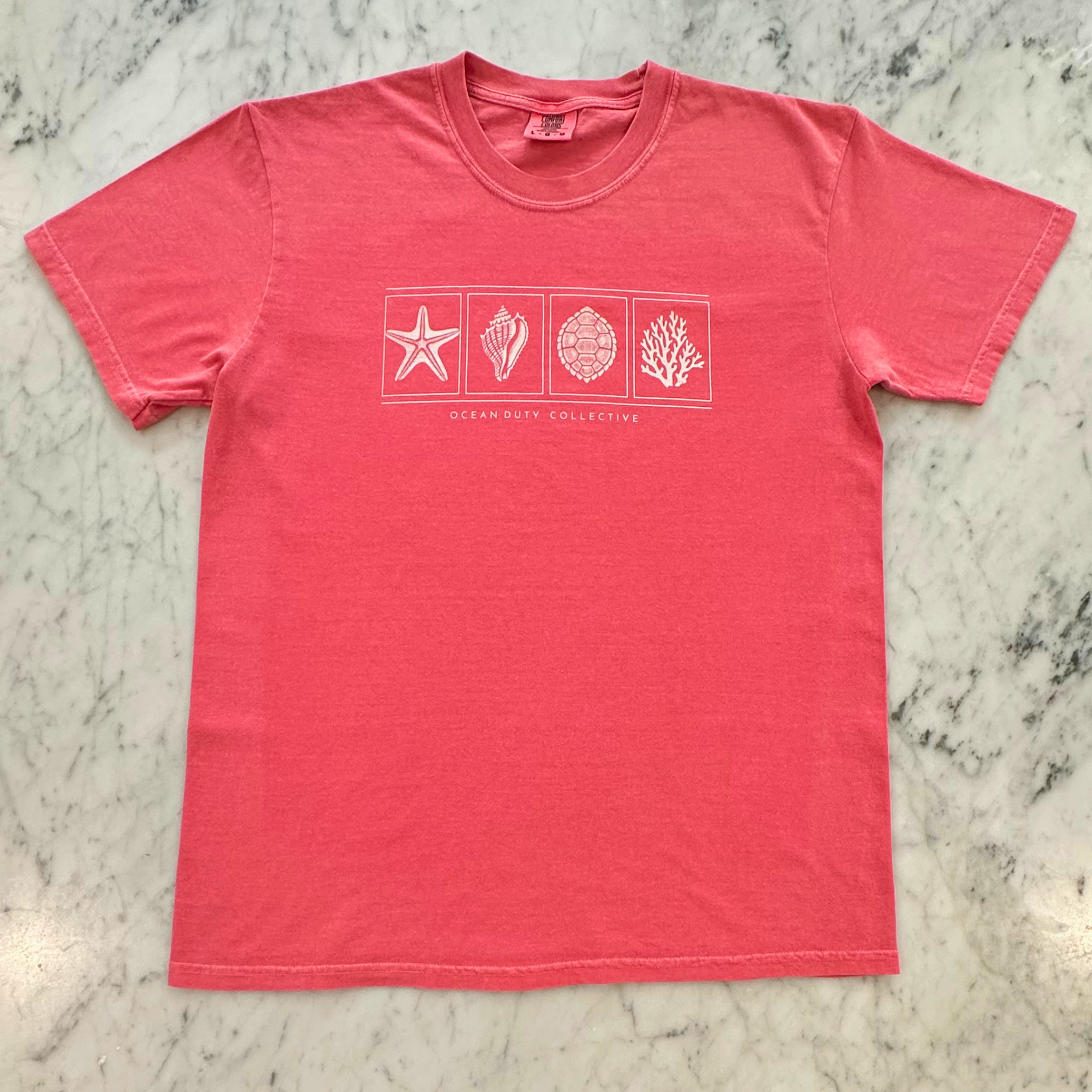 A coral pink short-sleeve t-shirt with a white graphic print of seashells and the words 'Ocean Way College' on the chest, laid flat on a gray and white marble surface.