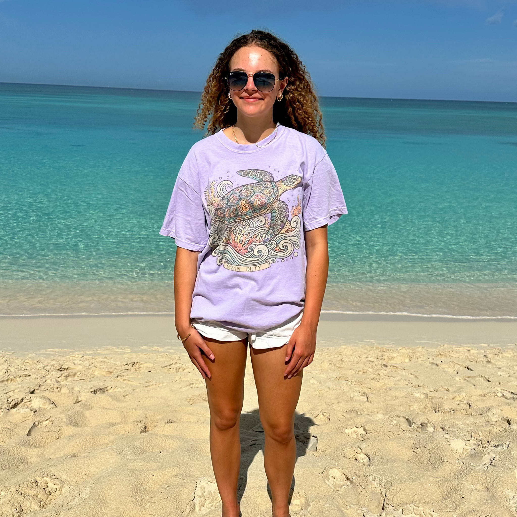 A woman stands on a sandy beach wearing a light purple graphic t-shirt with a sea turtle design, white shorts, and sunglasses, with the turquoise ocean and blue sky in the background.