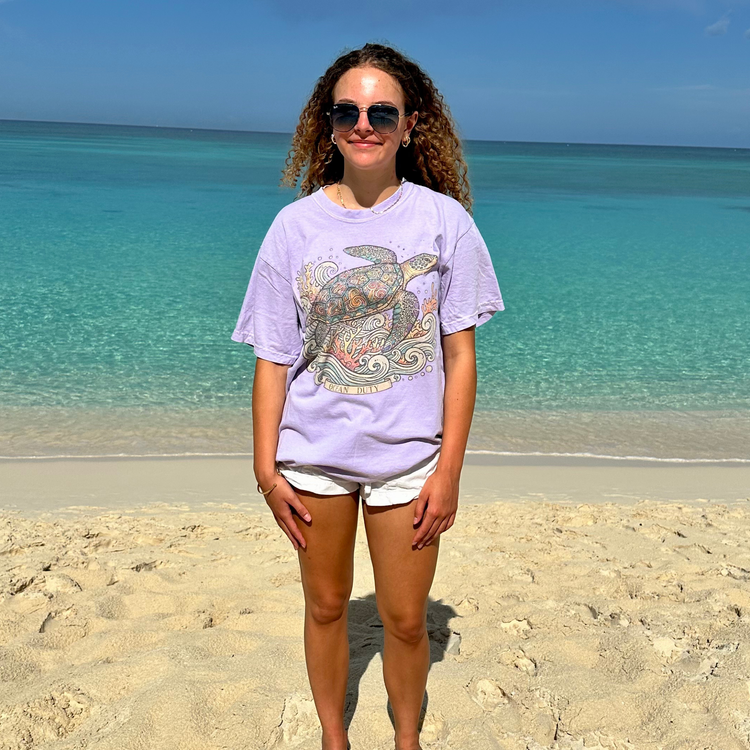 A woman stands on a sandy beach wearing a light purple graphic t-shirt with a sea turtle design, white shorts, and sunglasses, with the turquoise ocean and blue sky in the background.