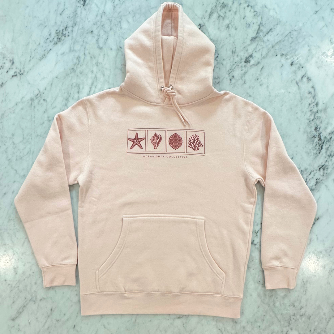 A light blush pink pullover hoodie with a front kangaroo pocket and a chest graphic showing four square icons (a starfish, a clam shell, a spiral shell, and coral) in a dusty rose color, laid flat on a light green marble surface. The brand name &