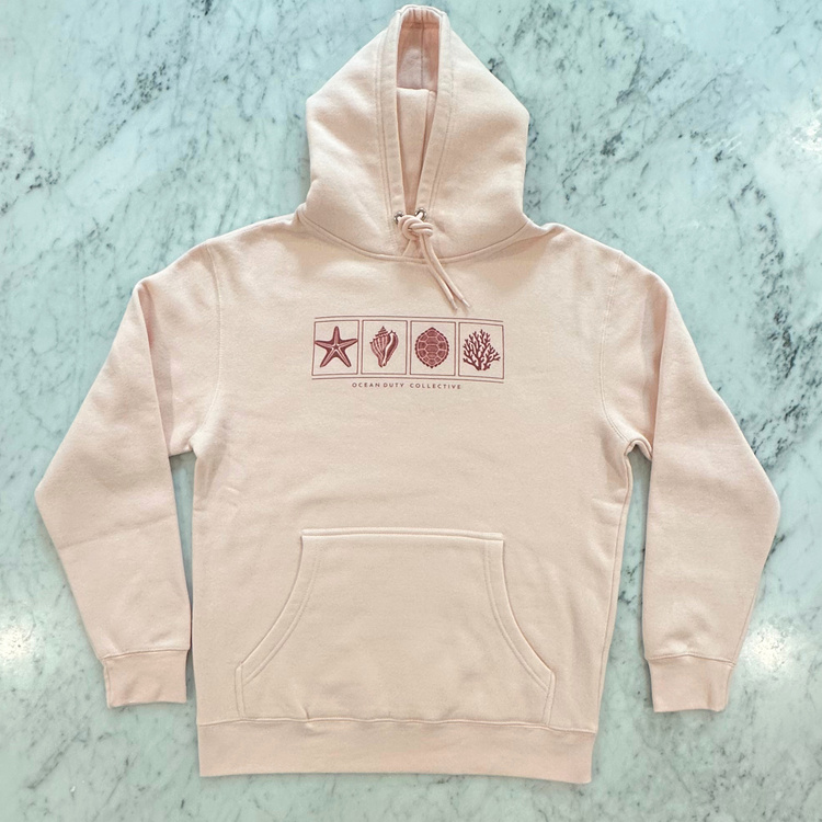 A light blush pink pullover hoodie with a front kangaroo pocket and a chest graphic showing four square icons (a starfish, a clam shell, a spiral shell, and coral) in a dusty rose color, laid flat on a light green marble surface. The brand name 'OCEAN DUTY COLLECTIVE' is printed below the graphic.