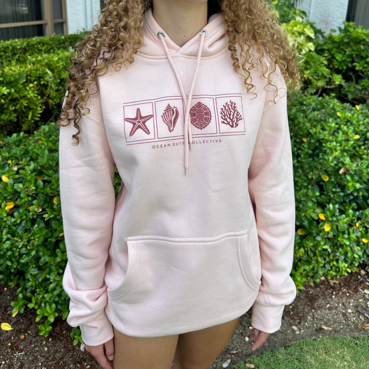 A person wearing a light pink hoodie with a dark pink ocean-themed graphic on the chest, standing outdoors in front of green bushes. The hoodie features a starfish, seashell, shell, and coral design with the text 'OCEAN DUTY COLLECTIVE' underneath.