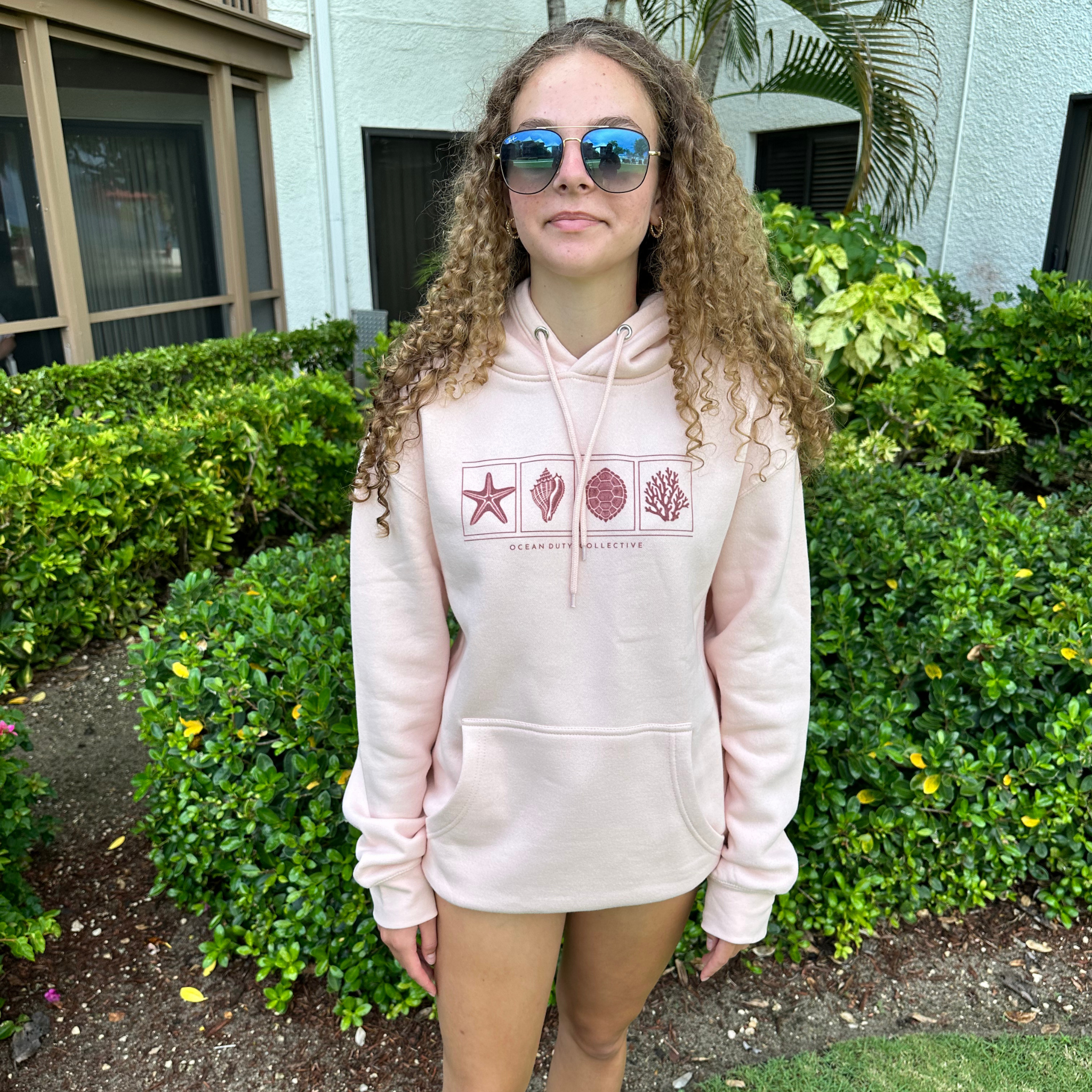 A woman with curly hair wearing a light pink hoodie with a seashell graphic print on the front, standing outdoors in front of green bushes and a building.