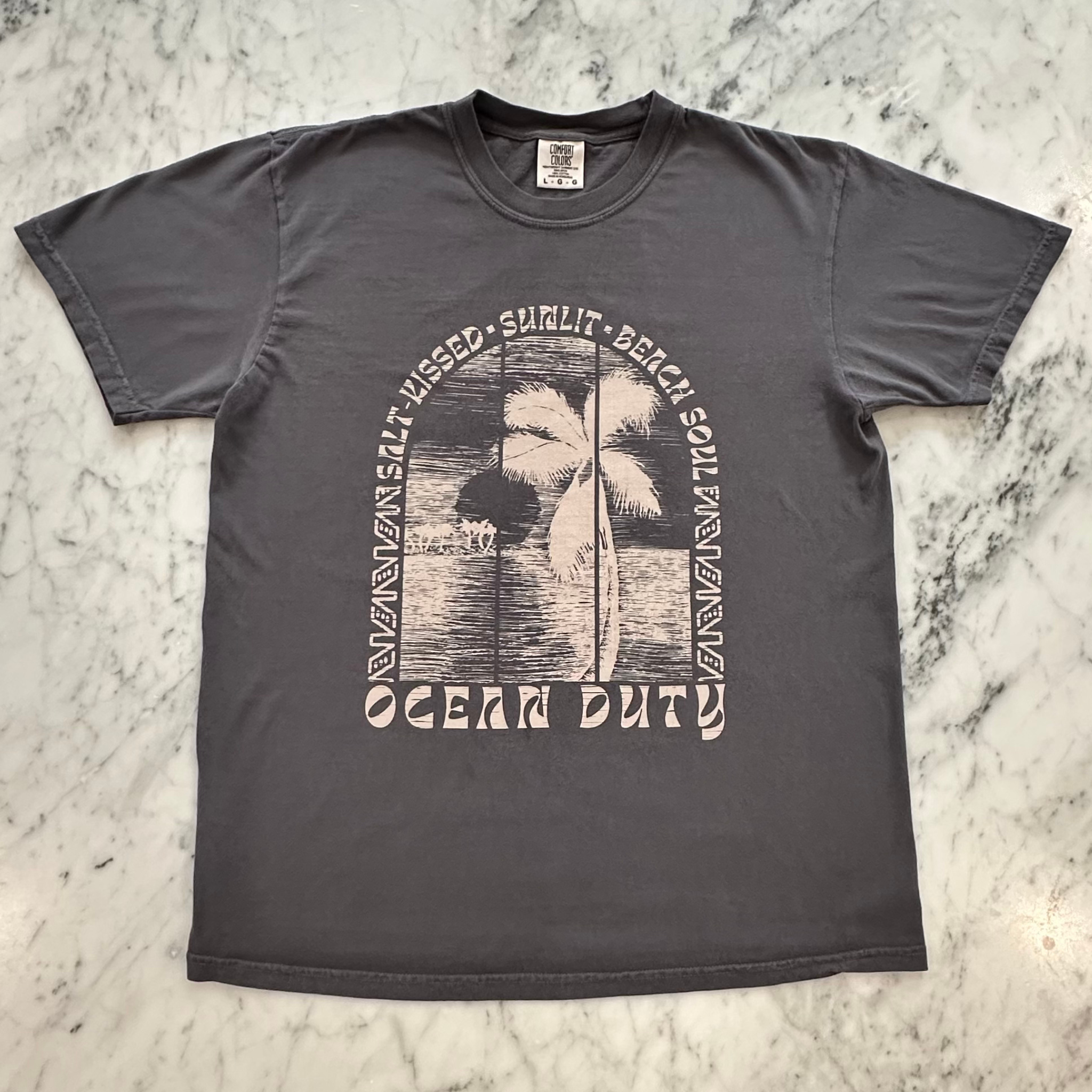 A dark gray graphic t-shirt laid flat on a white and gray marble surface. The shirt features a vintage-style graphic print in a light beige color showing a palm tree, a beach scene, and the words 'SALT KISSED, SUNLIT, BEACH SOUL' and 'OCEAN DUTY' printed below.