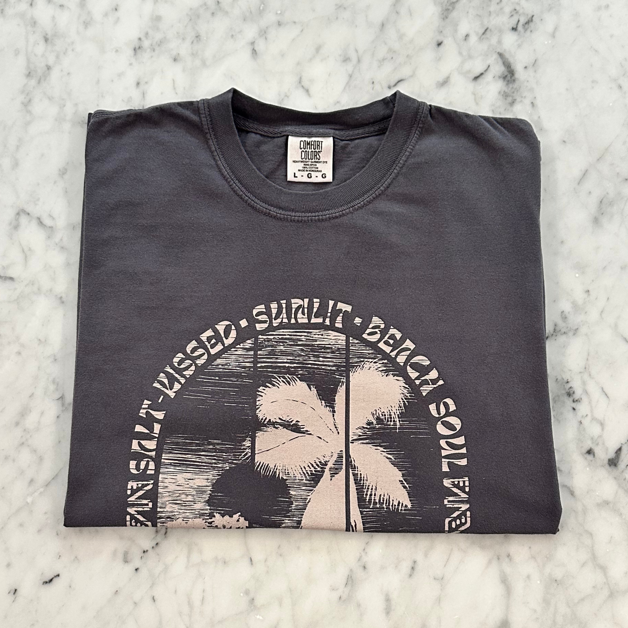 A folded dark charcoal gray t-shirt with a vintage-style palm tree graphic and the text 'SALT KISSED, SUNLIT, BEACH SOUL' on a white marble background.