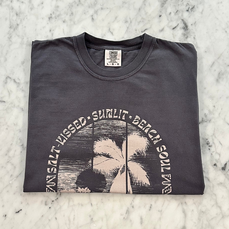 A folded dark charcoal gray t-shirt with a vintage-style palm tree graphic and the text 'SALT KISSED, SUNLIT, BEACH SOUL' on a white marble background.