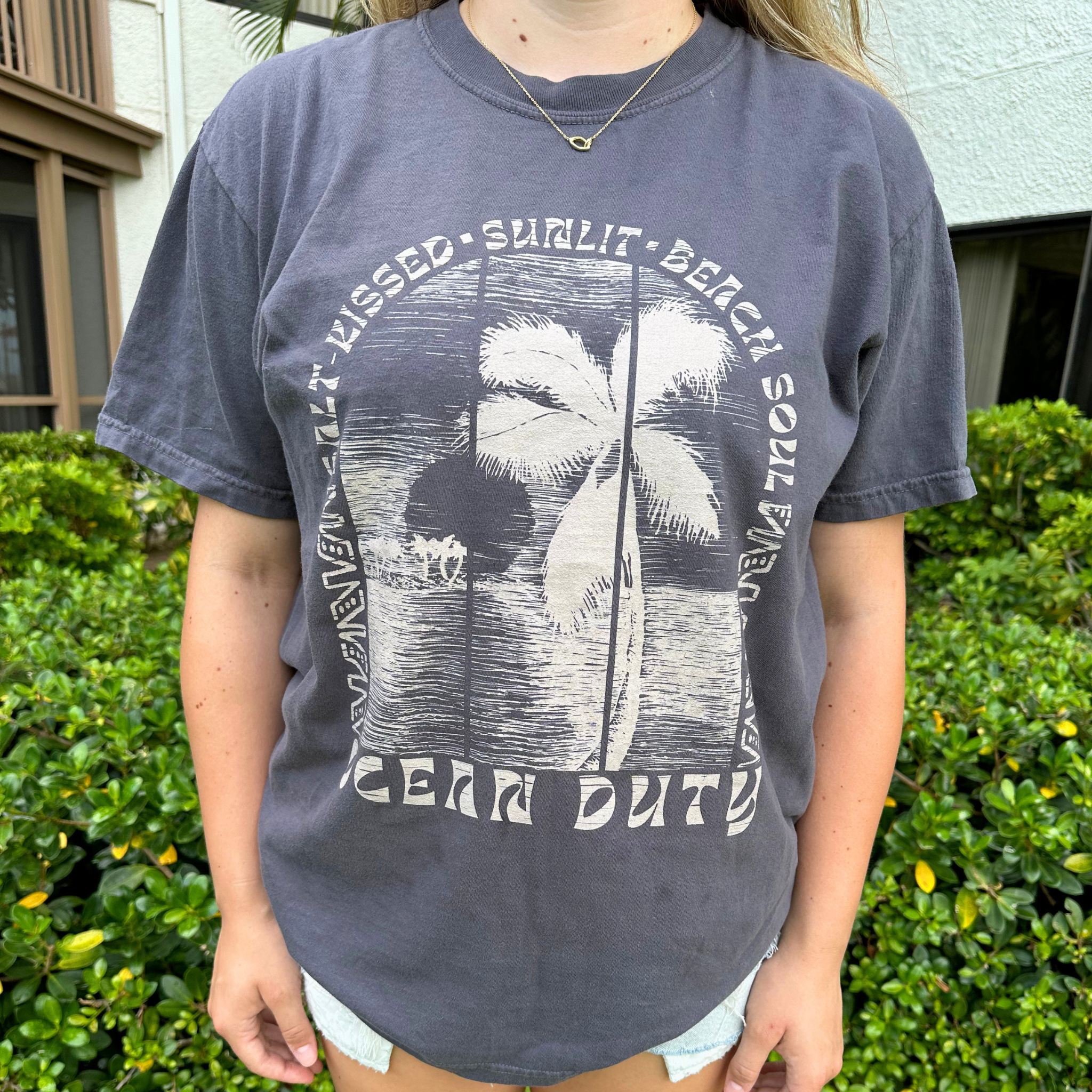 A person wearing a faded dark gray graphic t-shirt with a white palm tree design and text reading 'SALT KISSED, SUNLIT, BEACH SOUL'.