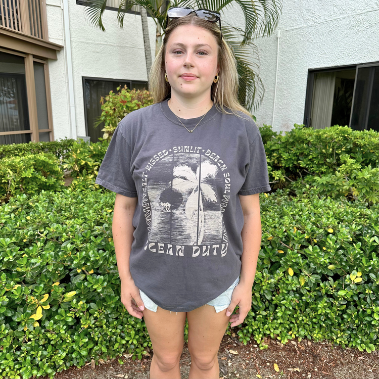 A young woman stands outdoors in front of a beach building and green hedges, wearing a dark gray graphic t-shirt with a palm tree design and the words 'SALT KISSED SUNLIT BEACH SOUL'.